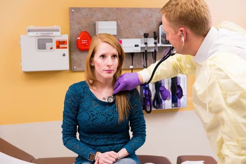 Una mujer se siente en el centro de cuidado médico. El doctor usa un estetoscopio para escucharle al corazón.