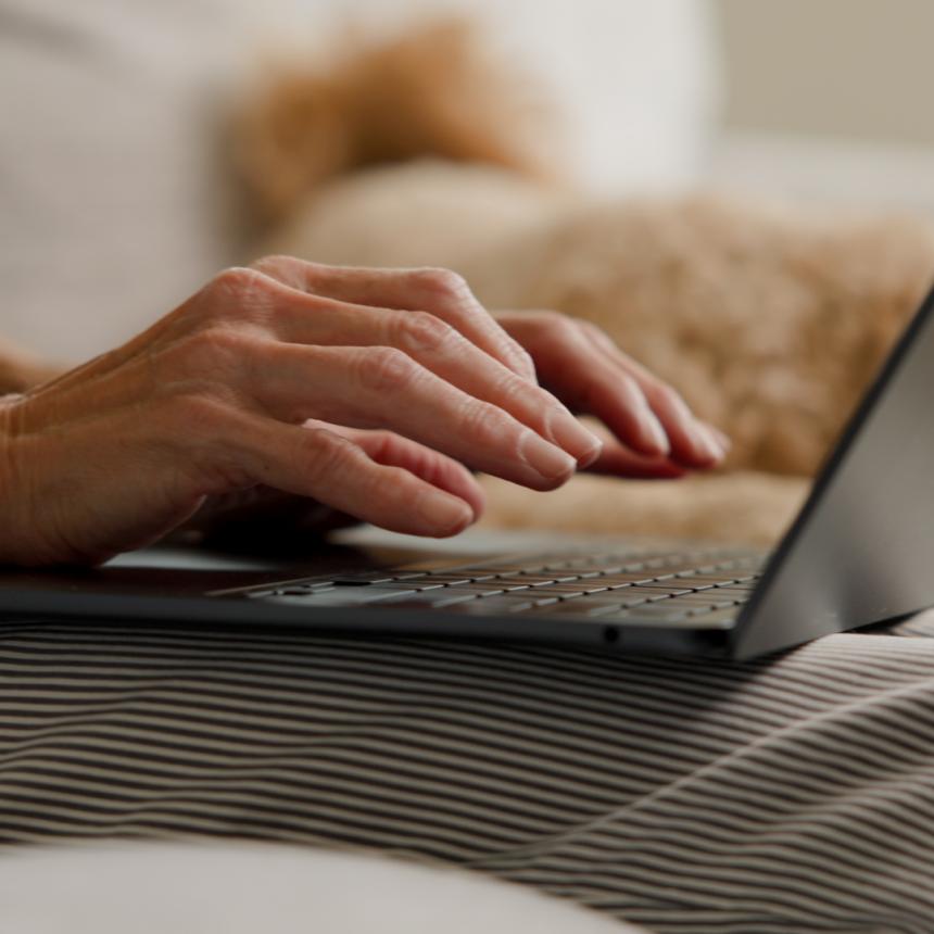 Una foto de manos sobre una computadora. La persona está sentada sobre un sofá, con un perro durmiendo al lado.