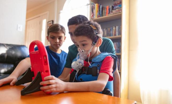 Un niño juega mientras hace sus tratamientos. Lleva un chaleco y usa un nebulizador.