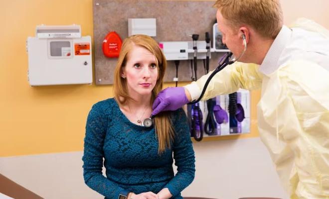 Una mujer se siente en el centro de cuidado médico. El doctor usa un estetoscopio para escucharle al corazón.